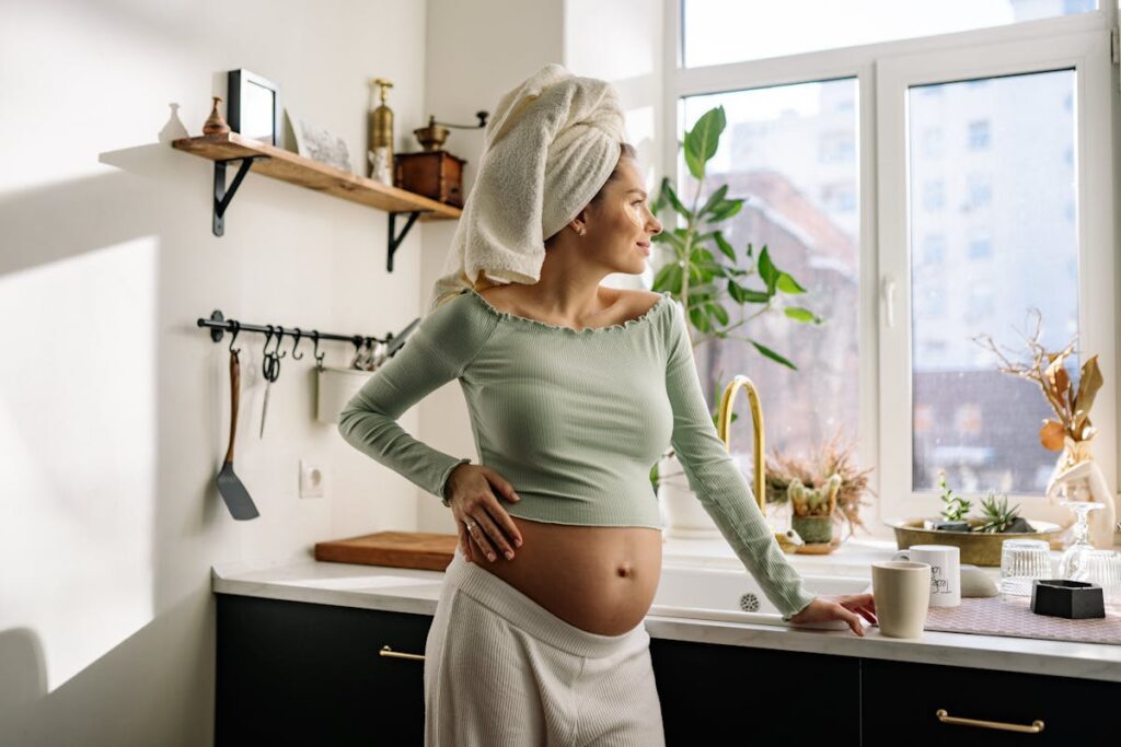 Future maman dans sa cuisine, main posee sur son ventre rond, lumiere naturelle du matin
